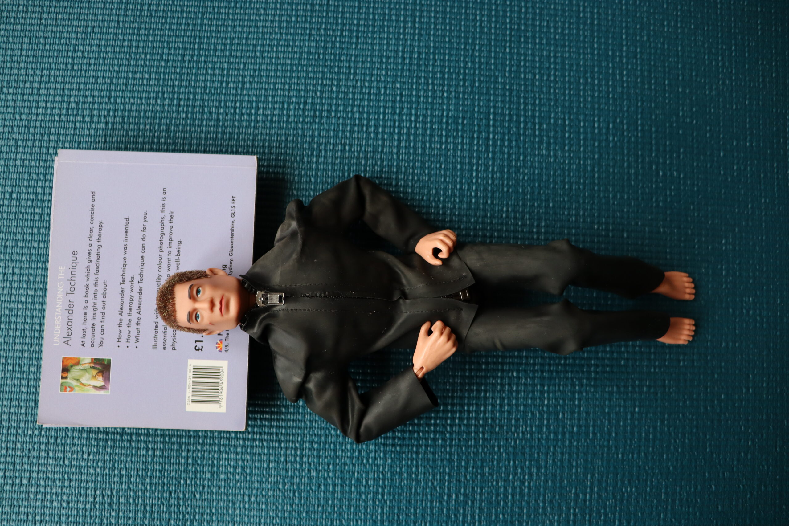 Photo of action man figure lying on a yoga mat with his head resting on some books, doing Alexander active rest