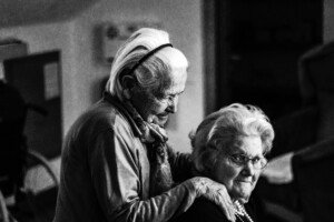 Photo of two older women, one with hands on the other's shoulders and both smiling